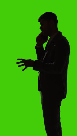 Vertikale-Videosilhouette-Eines-Geschäftsmannes,-Der-Vor-Grünem-Hintergrund-Mit-Dem-Mobiltelefon-Telefoniert